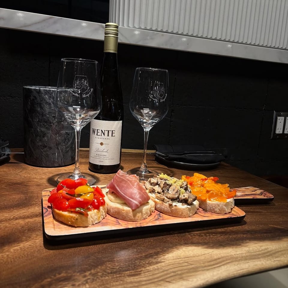 A bottle of Wente wine, two glasses, and a wooden board with four bruschetta appetizers.