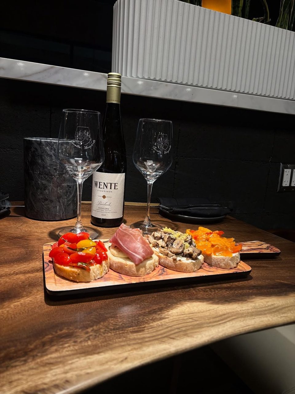 A bottle of Wente wine, two glasses, and a wooden board with four bruschetta appetizers.