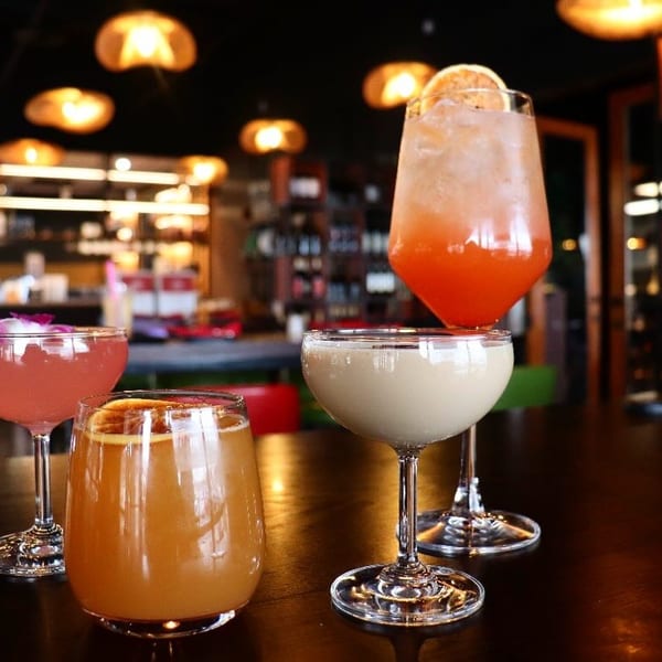Four colorful cocktails, including pink, orange, and cream-colored drinks, on a dark bar top.