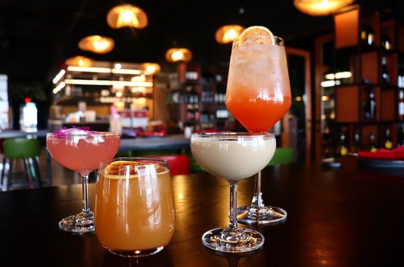 Four colorful cocktails, including pink, orange, and cream-colored drinks, on a dark bar top.