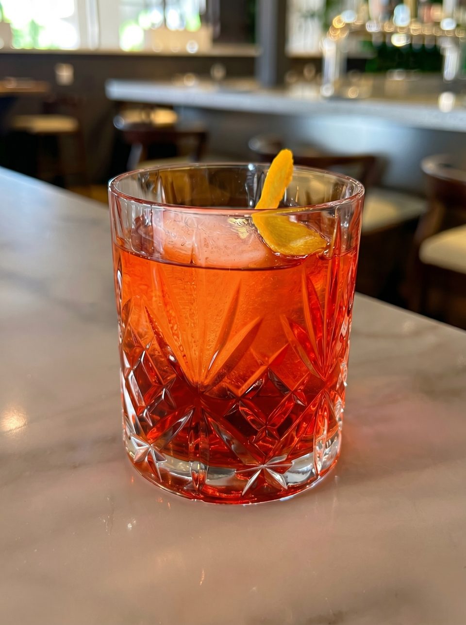 A vibrant red cocktail in a cut glass, with a large ice cube and orange peel, on a bar.