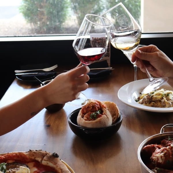 Two people clinking wine glasses over a table filled with Italian dishes like pasta and pizza.