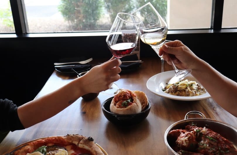 Two people clinking wine glasses over a table filled with Italian dishes like pasta and pizza.