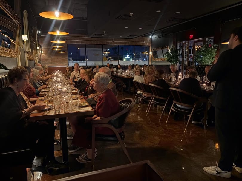 Diners seated at long tables in a dimly lit restaurant with warm overhead lighting and candles.