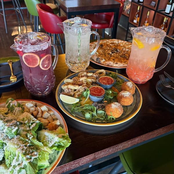 A restaurant table laden with pizza, salad, calamari, arancini, and three pitchers of colorful drinks.