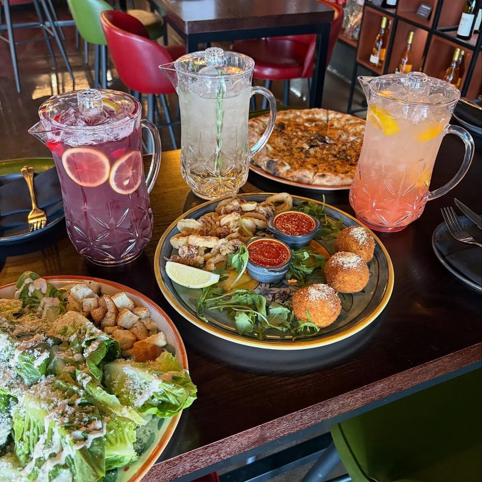 A restaurant table laden with pizza, salad, calamari, arancini, and three pitchers of colorful drinks.