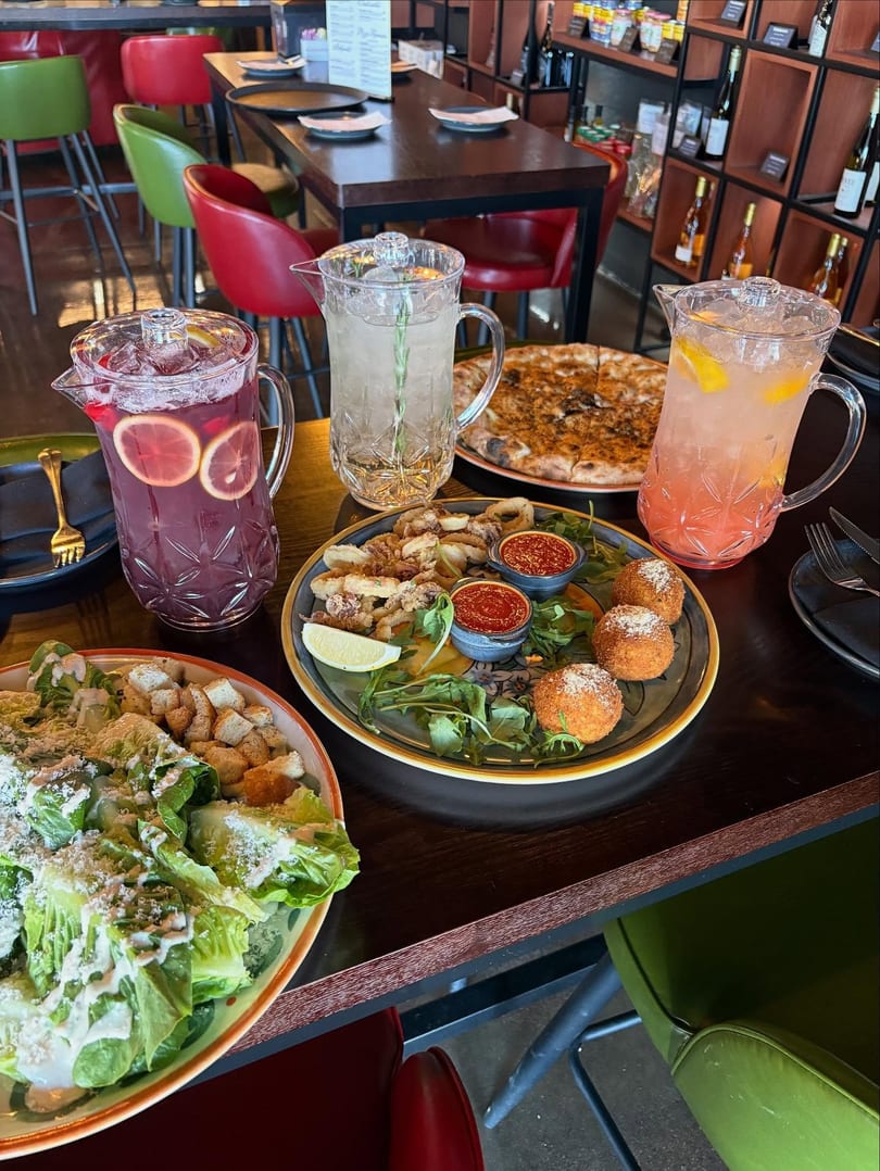 A restaurant table laden with pizza, salad, calamari, arancini, and three pitchers of colorful drinks.