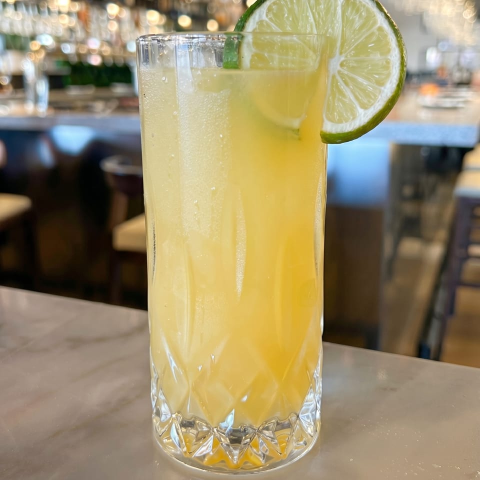 Tall glass of yellow cocktail with a lime slice on a marble bar, blurred lights in background.