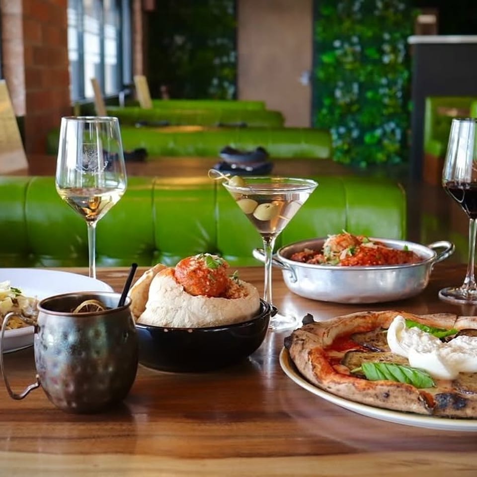 A table with various Italian dishes and drinks, including pizza, pasta, meatballs, and cocktails, in a restaurant.