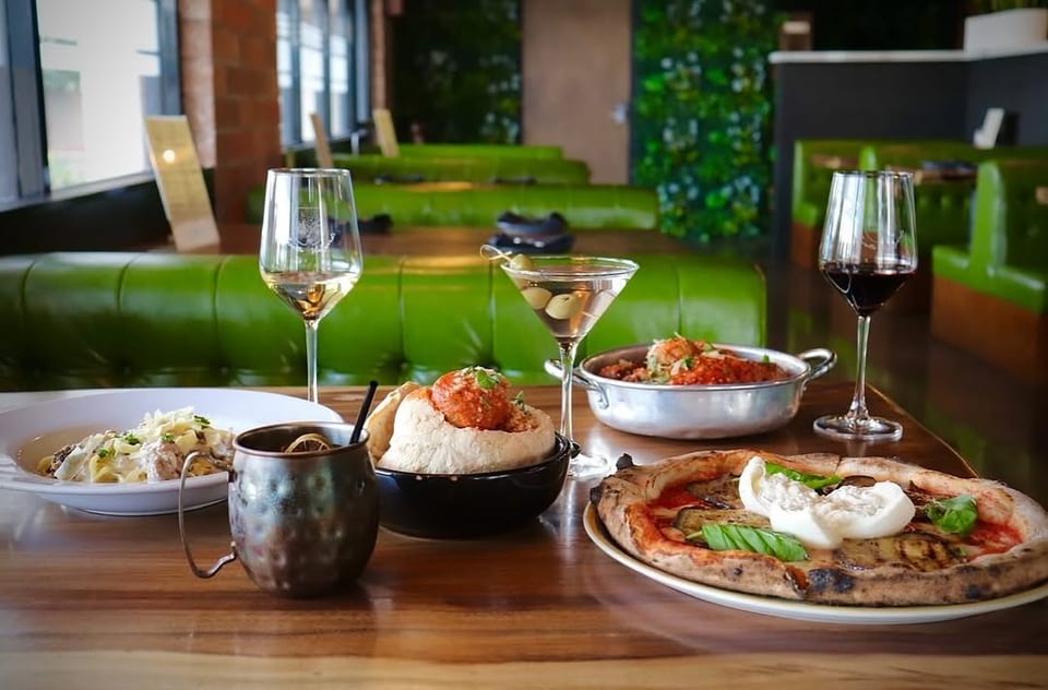 A table with various Italian dishes and drinks, including pizza, pasta, meatballs, and cocktails, in a restaurant.