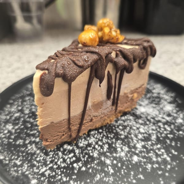 Slice of layered chocolate and caramel cheesecake with chocolate drizzle, nuts, and powdered sugar.