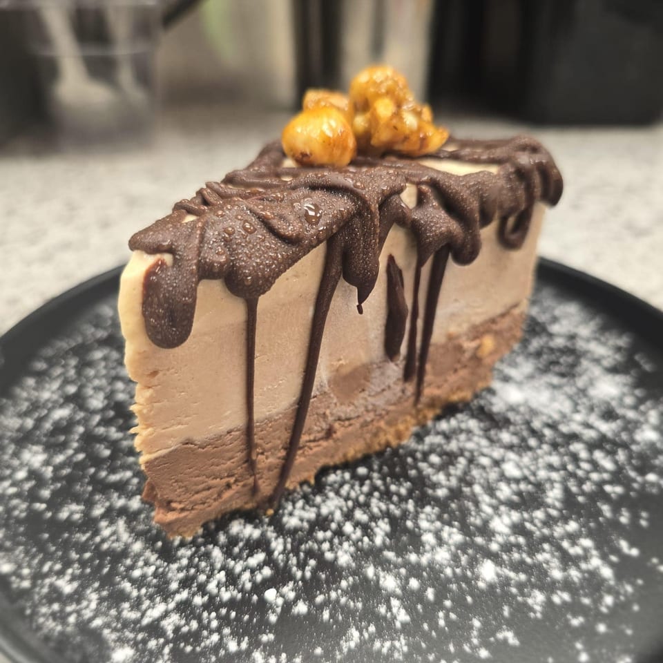 Slice of layered chocolate and caramel cheesecake with chocolate drizzle, nuts, and powdered sugar.