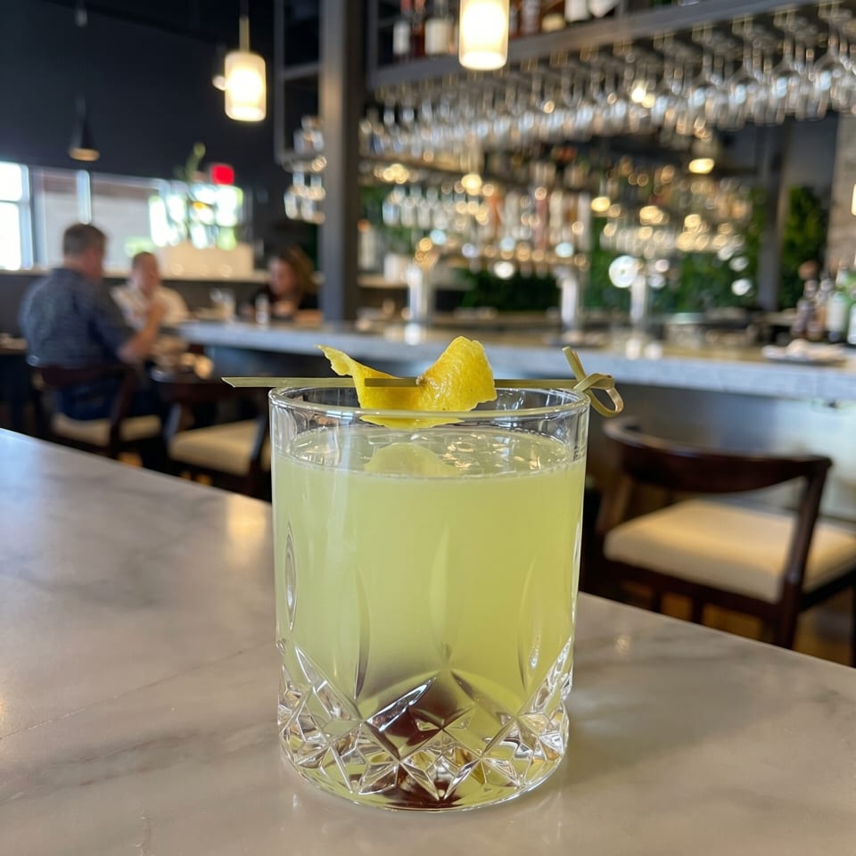 A yellow cocktail with a lemon peel garnish sits on a marble bar top in a restaurant.