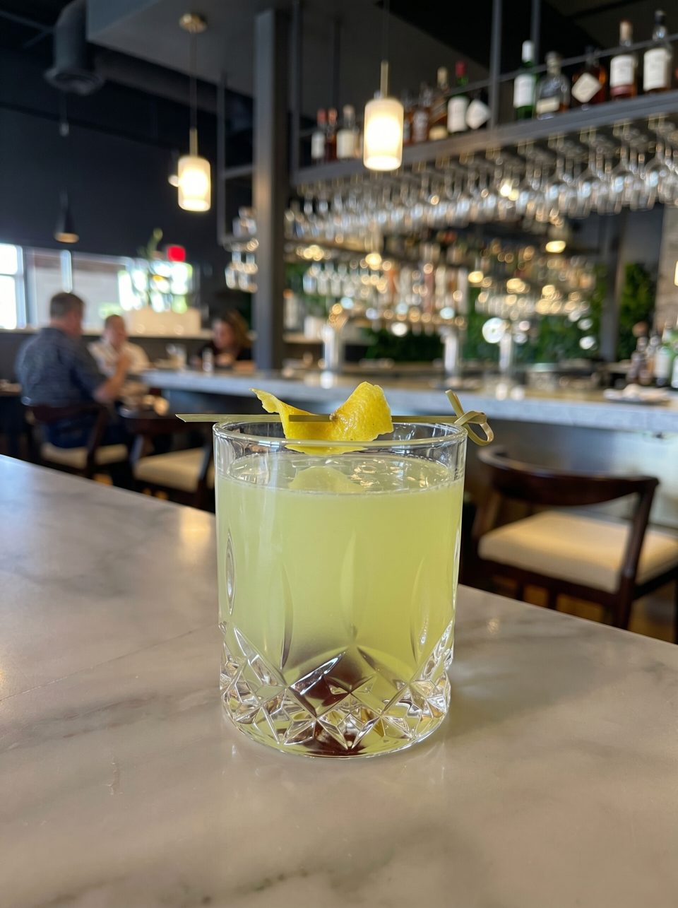 A yellow cocktail with a lemon peel garnish sits on a marble bar top in a restaurant.