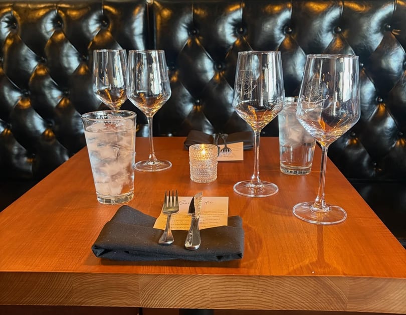 A set table with wine glasses, water glasses, silverware, and a lit candle against a dark tufted booth.