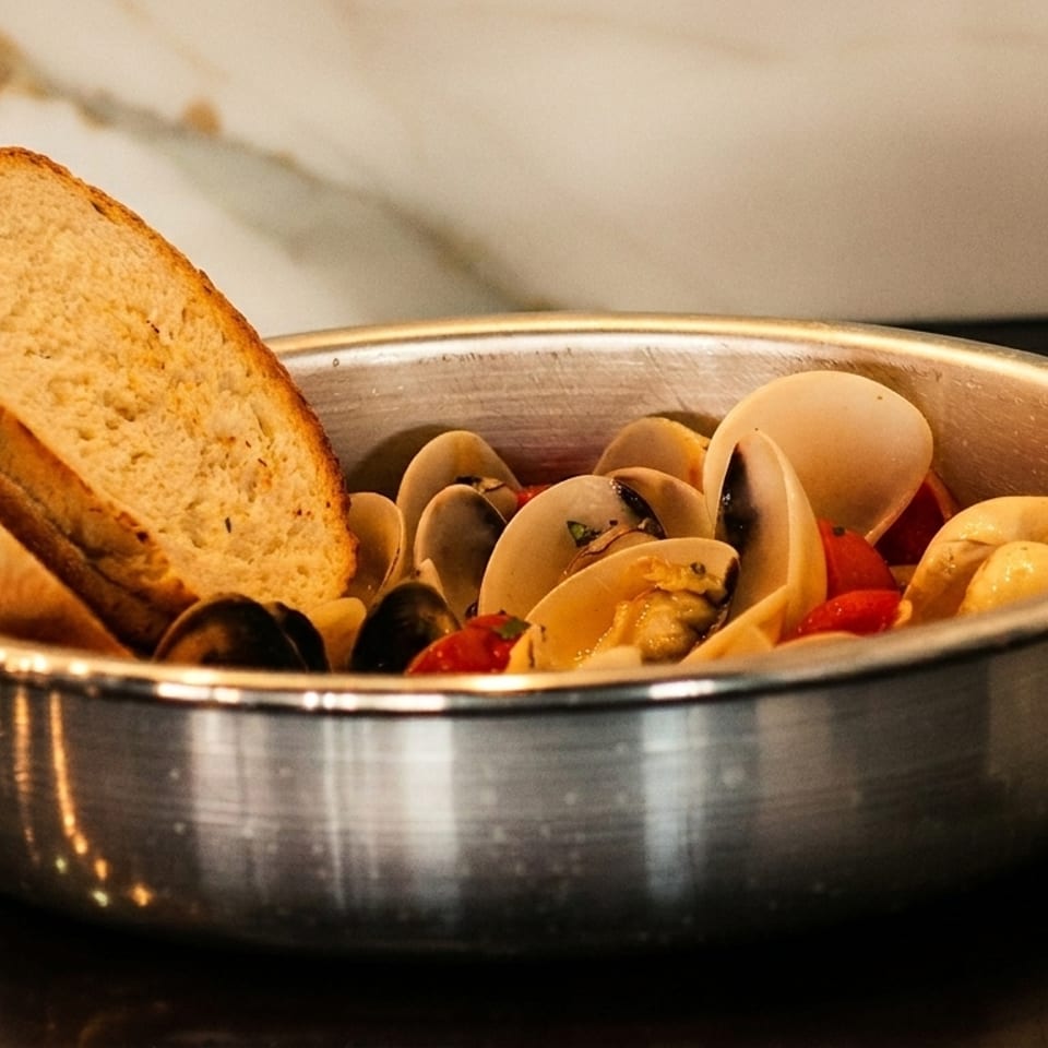 Steamed clams with cherry tomatoes and two slices of toasted bread in a metal pot.