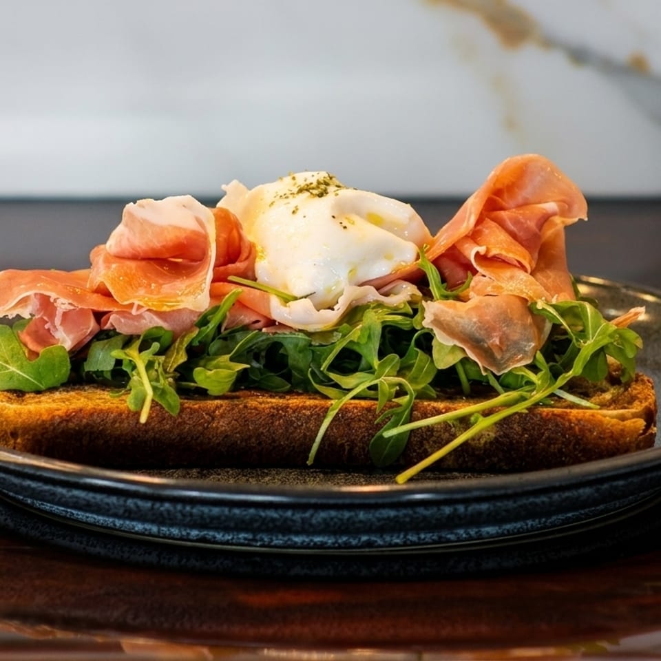 Toasted bread topped with prosciutto, arugula, and a poached egg on a dark plate.