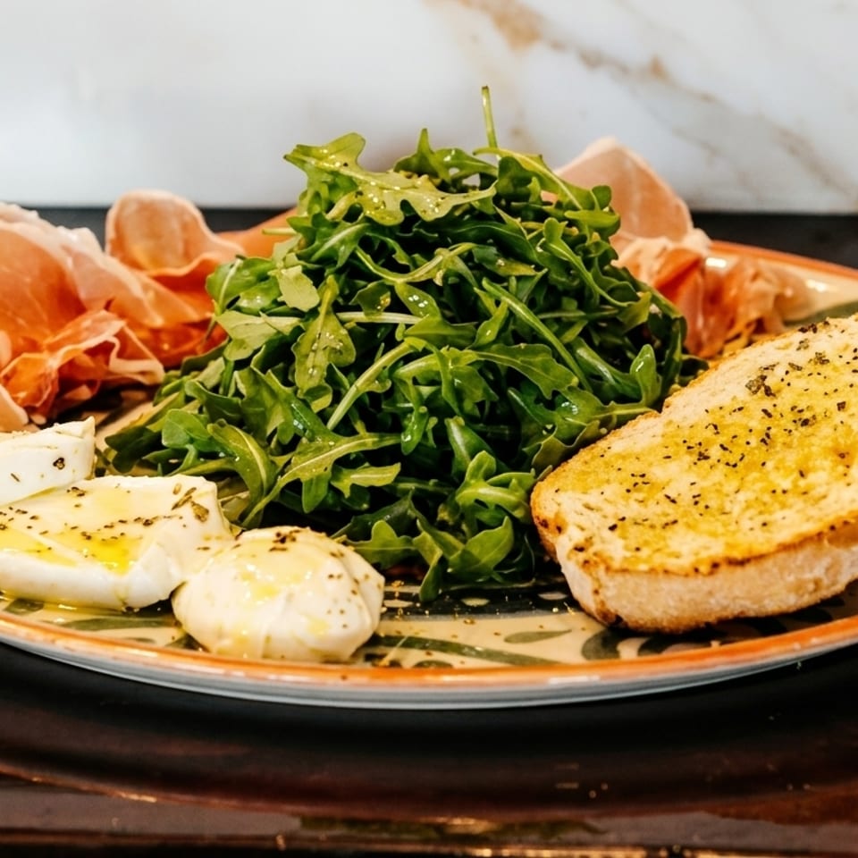 Appetizer plate with prosciutto, arugula, mozzarella, and garlic bread on a decorative plate.