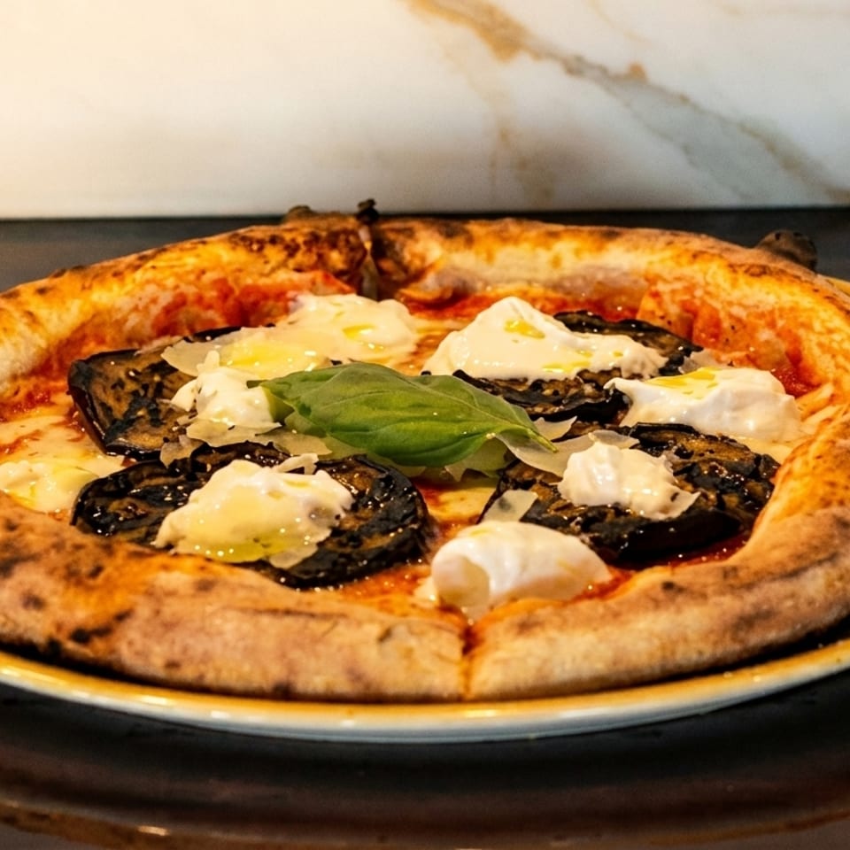 Neapolitan pizza with charred crust, eggplant, burrata, parmesan, and basil on a dark table.