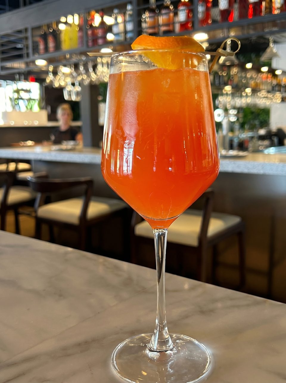 An orange cocktail with an orange slice garnish sits on a marble bar top.
